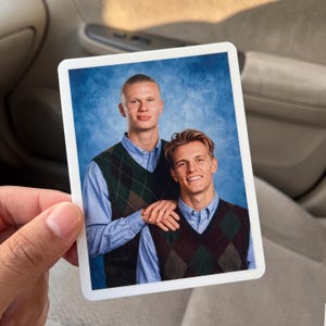 May include: A rectangular sticker featuring a portrait of two individuals wearing light blue button-down shirts under dark green and brown argyle sweater vests against a mottled blue background, with one individual resting a hand on the other's shoulder.