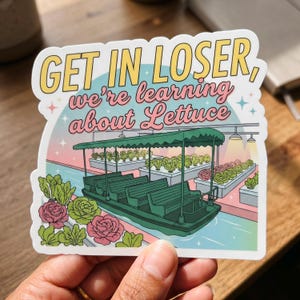 May include: A sticker with the text "GET IN LOSER, we're learning about Lettuce" in yellow and pink. The sticker features a green boat with benches, floating in a blue canal, surrounded by lettuce plants and pink roses.