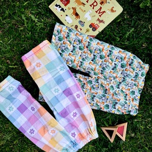May include: A children's book titled "FARM" with animal illustrations, alongside two pairs of patterned trousers and a wooden toy on green grass. One pair of trousers features a pastel plaid pattern with flower accents, the other has a jungle animal print.