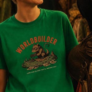 May include: A green short-sleeved t-shirt featuring a graphic of a creature wearing goggles and building a landscape. The text "WORLDBUILDER" appears in orange above the creature, with "LORE FOR PLACES YOU'LL NEVER VISIT" printed in white below.