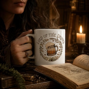 May include: A white ceramic mug featuring a brown graphic illustration of a frothy beer stein surrounded by a wreath of wheat and leaves, with the text "LORD OF THE DRINKS" arched above the design in a dark brown serif font.