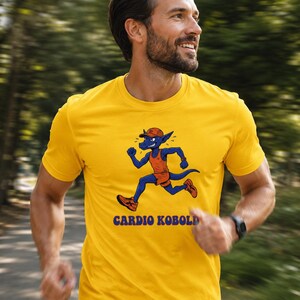 Puede incluir: Camiseta amarilla con un dibujo animado de una criatura azul corriendo, con un atuendo naranja y una gorra roja. El texto "CARDIO KOBOLD" está impreso debajo. La persona que lleva la camiseta está corriendo al aire libre.