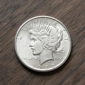 May include: A silver Peace dollar coin from 1925, featuring a profile of Lady Liberty with rays emanating from her crown. The coin is inscribed with "IN GOD WE TRUST" and "LIBERTY". The coin is on a dark wood surface.