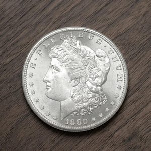 Może przedstawiać: Srebrna moneta Morgan Dollar z 1880 roku, przedstawiająca profil Lady Liberty i napis "E PLURIBUS UNUM". Moneta jest umieszczona na tle ciemnego drewna.