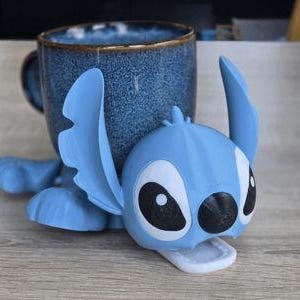 May include: A blue ceramic mug with a textured surface and a gold rim, resting on a light blue Stitch figure. The Stitch figure has large black and white eyes and a white mouth. The mug and figure are on a wooden surface.
