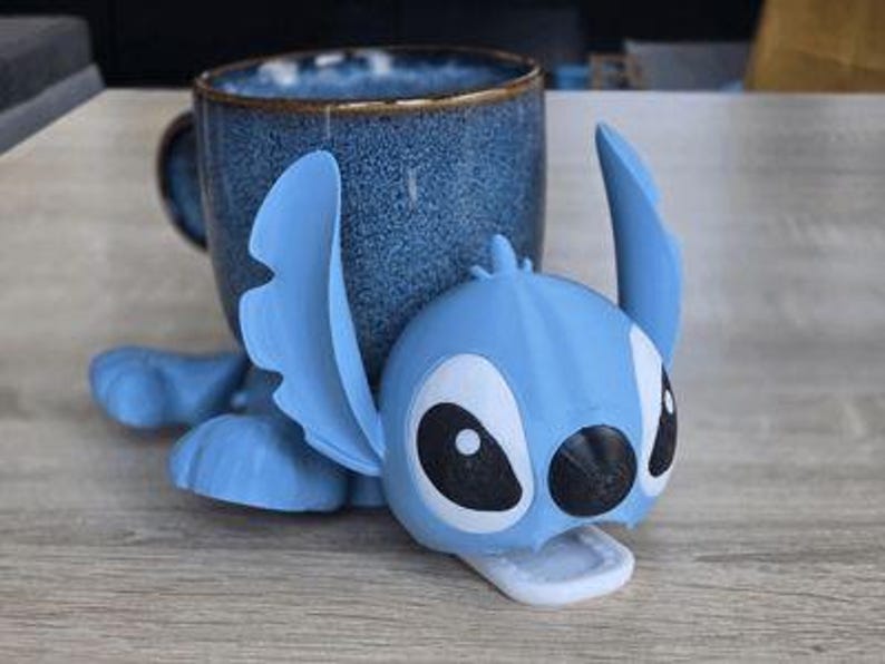 May include: A blue ceramic mug with a textured surface and a gold rim, resting on a light blue Stitch figure. The Stitch figure has large black and white eyes and a white mouth. The mug and figure are on a wooden surface.