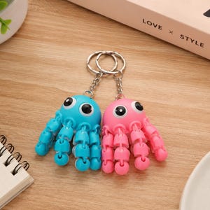 May include: Two octopus keyrings, one blue and one pink, with large googly eyes and articulated tentacles. The keyrings are made of plastic and attached to silver key rings. The background includes a wooden surface, a notebook, and a box with the words "LOVE x STYLE x LIFE."