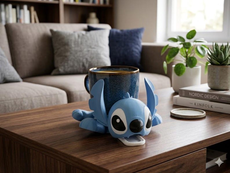 May include: A blue ceramic mug with a gold rim rests in a light blue Stitch-shaped holder on a wooden coffee table. The mug is filled with a dark liquid. Decorative plants and books are in the background.