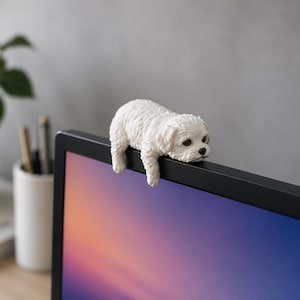 Puede incluir: Una figura de perro blanco y esponjoso posada en el borde superior de un monitor de ordenador. El perro pequeño tiene un pelaje texturizado y está tumbado, con las patas delanteras colgando. El monitor muestra un degradado de colores.