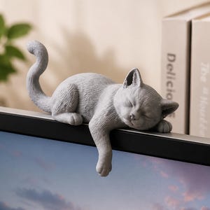 May include: A gray cat figurine is resting on a black surface. The cat is curled up with its eyes closed, and one paw is dangling over the edge. The figurine is detailed and appears to be made of stone or a similar material.