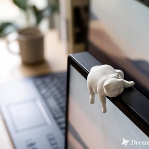 May include: A white ceramic cat figurine, designed to hang on the edge of a computer monitor. The cat is in a relaxed pose, with its paws dangling. The background includes a laptop, a mug, and a plant.