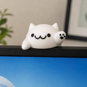 May include: A white cat-shaped desk accessory with a friendly expression. The cat has black eyes, a smiling mouth, and a paw raised in a wave. The accessory is placed on top of a black computer monitor.