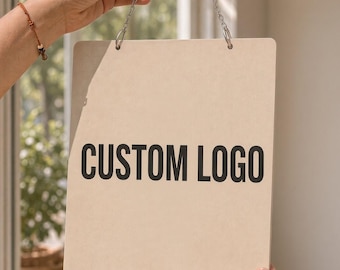 Może przedstawiać: Prostokątny, jasnobeżowy szyld z czarnym napisem CUSTOM LOGO. Szyld jest zawieszony na dwóch srebrnych metalowych łańcuszkach przymocowanych do górnych rogów, co zapewnia przejrzystą powierzchnię do celów reklamowych.