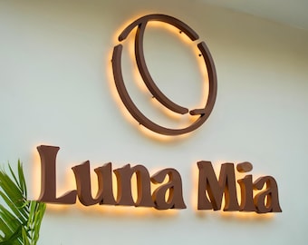 May include: A wall-mounted sign featuring the text Luna Mia in dark brown, dimensional lettering. Above the text is a circular, crescent-shaped logo. Both the letters and the logo are backlit with a warm, glowing light against a plain wall.