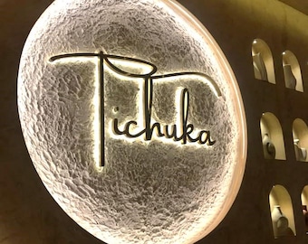 May include: A circular wall sign featuring the word Tichuka in elegant gold script lettering, mounted on a textured, off-white, moon-like surface with warm backlighting. To the right, a wall contains several arched niches displaying vases.