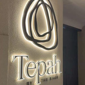 Może przedstawiać: Naścienny szyld z ciemnym, abstrakcyjnym, okrągłym logo nad napisem "Tepah" i "BY THE BAGH" wykonanym czcionką szeryfową. Elementy są podświetlone ciepłym białym światłem, tworząc efekt poświaty na teksturowanej powierzchni ściany.