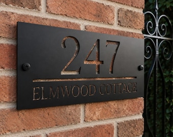 May include: A rectangular black metal house sign featuring the laser-cut number 247 and the text ELMWOOD COTTAGE engraved below. The sign is secured to a brick wall with two visible black screws on the left and right sides of the metal plate.
