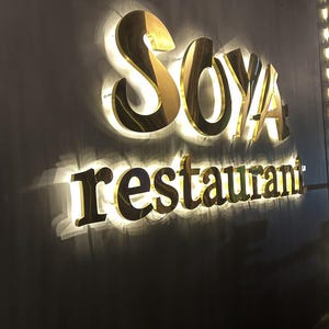 Może przedstawiać: Tr&oacute;jwymiarowy szyld ścienny z napisem SOYA wykonanym z dużych, złotych wielkich liter, umieszczonym nad słowem restaurant zapisanym mniejszymi, złotymi małymi literami, oba elementy podświetlone jasnym efektem podświetlenia tła.