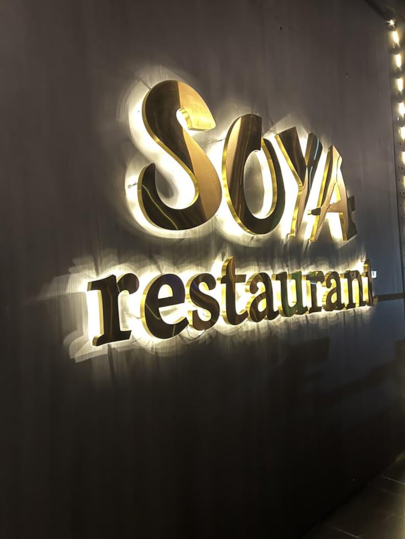 Może przedstawiać: Tr&oacute;jwymiarowy szyld ścienny z napisem SOYA wykonanym z dużych, złotych wielkich liter, umieszczonym nad słowem restaurant zapisanym mniejszymi, złotymi małymi literami, oba elementy podświetlone jasnym efektem podświetlenia tła.