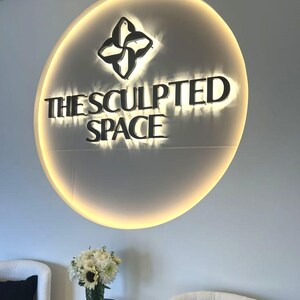 Może przedstawiać: Okrągły znak ścienny z czarnym napisem THE SCULPTED SPACE oraz logo z czterema płatkami. Znak jest podświetlony ciepłym światłem, tworząc efekt aureoli na ścianie. Poniżej znajduje się bukiet białych kwiat&oacute;w.