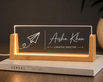 May include: A rectangular clear acrylic desk sign with warm LED illumination, held by a light wood base. The sign features a white etched paper airplane graphic with a small heart trail, the name Aisha Khan, and the title CREATIVE DIRECTOR.