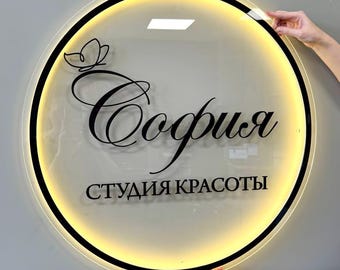 May include: A circular, transparent acrylic sign with a black border and internal warm yellow LED backlighting. The sign features a black butterfly graphic and the Cyrillic text "София" in a script font, followed by "СТУДИЯ КРАСОТЫ" in a sans-serif font.
