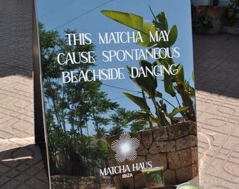 May include: A silver mirrored A-frame sign featuring the white text WARNING THIS MATCHA MAY CAUSE SPONTANEOUS BEACHSIDE DANCING and the logo MATCHA HAUS IBIZA. A clear plastic cup containing a layered green and red matcha drink sits in front.
