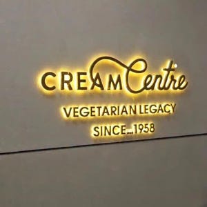 Może przedstawiać: Ścienny szyld z napisem Cream Centre stylizowaną złotą czcionką z ż&oacute;łtym podświetleniem. Poniżej tekst VEGETARIAN LEGACY oraz SINCE 1958 wyświetlane są w tym samym stylu podświetlanych złotych liter.