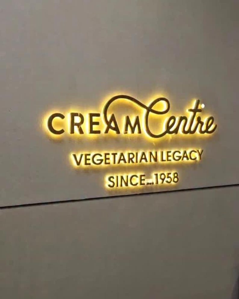 Może przedstawiać: Ścienny szyld z napisem Cream Centre stylizowaną złotą czcionką z ż&oacute;łtym podświetleniem. Poniżej tekst VEGETARIAN LEGACY oraz SINCE 1958 wyświetlane są w tym samym stylu podświetlanych złotych liter.