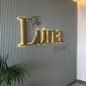 Może przedstawiać: Ścienny szyld ze złotym napisem The Luna CLUB. Litery są podświetlone ciepłym światłem i zamontowane na jasnoszarym panelu z pionowymi żłobieniami. W lewym dolnym rogu widoczna jest zielona roślina doniczkowa.
