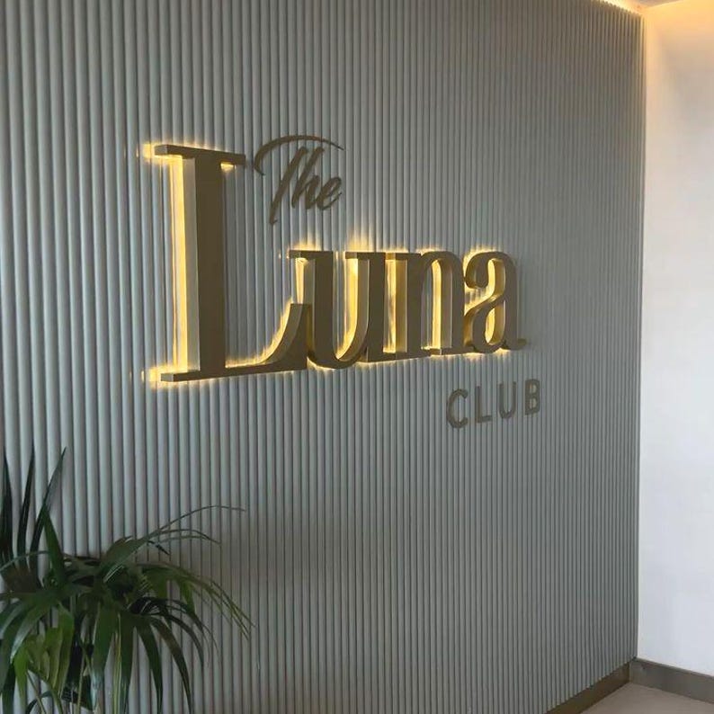 Może przedstawiać: Ścienny szyld ze złotym napisem The Luna CLUB. Litery są podświetlone ciepłym światłem i zamontowane na jasnoszarym panelu z pionowymi żłobieniami. W lewym dolnym rogu widoczna jest zielona roślina doniczkowa.