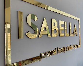 May include: A rectangular wall sign featuring a gold mirrored finish with the name ISABELLA in large capital letters and the text personal beauty studio & spa in smaller lowercase letters, all enclosed within a thin gold mirrored rectangular frame.