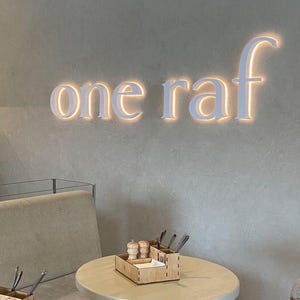 Może przedstawiać: Ściana z podświetlanym białym napisem "one raf" umieszczonym na teksturowanej szarej powierzchni. Poniżej znajduje się okrągły st&oacute;ł z drewnianym organizerem zawierającym solniczkę, pieprzniczkę i metalowe sztućce oraz krzesła.