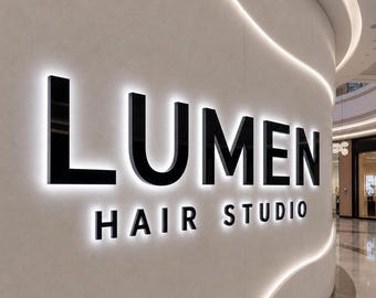 May include: A wall-mounted sign featuring the black text LUMEN HAIR STUDIO in a bold, sans-serif font. The letters are backlit with a bright white glow, creating a halo effect against the light-colored, curved wall surface of the interior space.