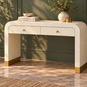 May include: A cream-colored console table with a textured finish, featuring two drawers with small gold knobs and curved side panels. The base of each leg is accented with a gold metal cap. Two books titled The Kinfolk Home are stacked on top.