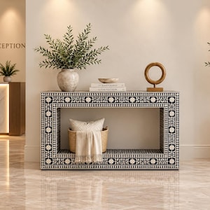 Peut inclure: Une console rectangulaire ornée d'un motif floral géométrique noir et blanc. La table présente un centre évidé contenant un panier tressé avec un textile beige. Un vase avec de la verdure, deux livres et une sculpture circulaire sont posés dessus.