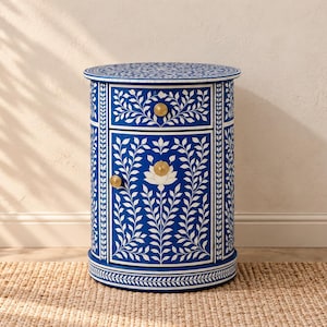 May include: A cylindrical bedside table featuring a deep blue finish with intricate white floral and vine inlays. The piece includes one top drawer and one lower cabinet door, each accented with small, round, gold-toned metal knobs for hardware.