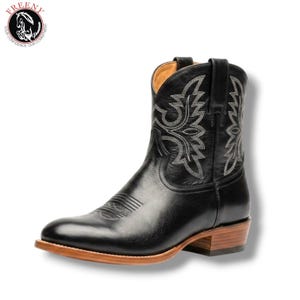 Puede incluir: Una bota vaquera de cuero negro con costuras decorativas blancas en la caña y la puntera. La bota tiene un forro interior color canela, un tacón de madera apilada y una suela marrón claro. Se observa un logotipo circular con un caballo.