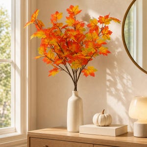 May include: A tall, slender white ceramic vase holds a large arrangement of artificial maple branches with vibrant autumn-coloured leaves in shades of red, orange, and yellow. The branches are displayed upright, creating a full, leafy appearance.