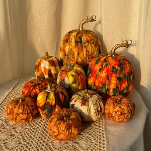May include: A collection of nine fabric pumpkins in various sizes, featuring assorted autumn-themed patterns including orange, black, and white prints, each topped with a metallic gold stem, arranged together on a textured white surface.