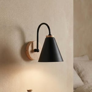 Può includere: Una lampada da parete con paralume conico nero opaco fissato a un braccio nero curvo e una base circolare in legno chiaro. La lampada è posizionata sopra un cassetto sospeso in legno con una pila di libri e un piccolo vaso in ceramica.