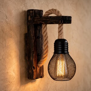 May include: A wall-mounted light fitting featuring a dark, distressed wooden beam bracket and a thick, natural-coloured jute rope that suspends a black metal mesh cage housing a glowing Edison-style light bulb with visible vertical filaments.