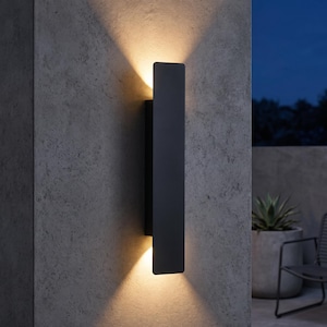 Pode incluir: Uma luminária de parede retangular preta fosca montada verticalmente em uma superfície cinza texturizada. A luminária emite um feixe de luz quente e focado pelas bordas superior e inferior, criando dois padrões de luz triangulares.