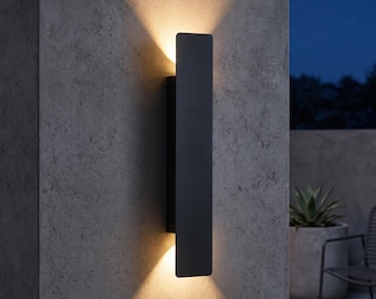 May include: A rectangular matte black wall sconce mounted vertically on a textured gray surface. The fixture emits a warm, focused beam of light from both the top and bottom edges, creating two distinct triangular light patterns on the wall.