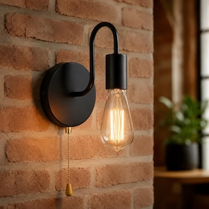 Pode incluir: Um candeeiro de parede preto mate com braço curvo, casquilho cilíndrico e uma lâmpada Edison em forma de gota. Uma corrente de puxar com extremidade dourada pende do suporte circular de parede, servindo como mecanismo de controlo.