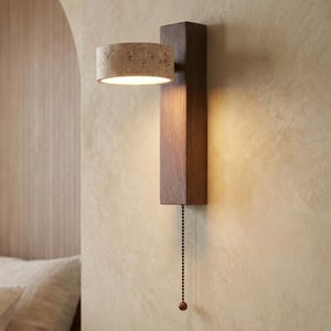 Peut inclure: Applique murale moderne avec une base rectangulaire en bois foncé et un abat-jour cylindrique. L'abat-jour est de couleur beige clair avec une surface texturée. Une chaîne pend de la base. L'applique est fixée sur un mur beige clair.