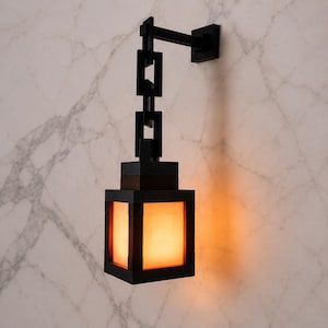 Peut inclure: Une lanterne murale noire dotée d'un cadre carré et d'une lumière orange chaude et éclatante. Le luminaire est suspendu par une chaîne verticale composée de trois maillons noirs rectangulaires fixés à un bras horizontal sur une plaque murale.