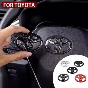 Op de afbeelding: Zwarte en koolstofvezel Toyota embleem overlay. De afbeelding toont een hand die een koolstofvezel embleem vasthoudt naast een stuurwiel met een chroom embleem. Extra emblemen in zilver, zwart en rood worden ook getoond. De tekst "FOR TOYOTA" staat linksboven.