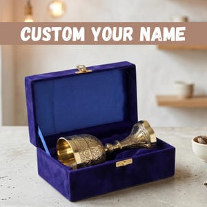 May include: A gold-colored metal chalice with intricate embossed patterns resting inside a rectangular, deep purple velvet-lined presentation box featuring a gold-tone metal latch. The text CUSTOM YOUR NAME is displayed above the object.