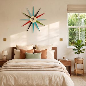 May include: A starburst wall clock featuring a central cream-colored circular face with black hands and twelve radiating pointed spikes in alternating colors of red, black, light blue, and green, mounted on a plain wall above a bed.